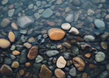 Clear river water over smooth natural pebbles with soft ripples and earthy tones creating calm relaxing nature aesthetic wallpaper