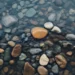 Clear river water over smooth natural pebbles with soft ripples and earthy tones creating calm relaxing nature aesthetic wallpaper