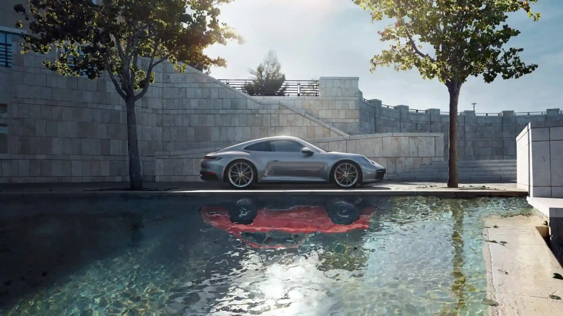 Silver Porsche 911 Carrera sports car parked beside a reflective pool with modern stone architecture, green trees, and warm sunlight in a Full HD live wallpaper.
